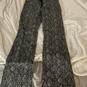 Grå bootcut byxor med ormmönster - Säljer ett par grå bootcut byxor med snyggt ormmönster och snörning i midjan. Byxorna har två stora bakfickor. Storlek 42 från monki. Nypris 600kr
