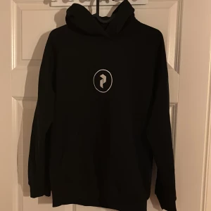 Svart hoodie från Peak Performance - Svart hoodie från Peak Performance med stor logga i vitt på ryggen och mindre logga framtill. Klassisk huva och känguruficka. Perfekt för chill eller när du vill ha en clean look. Använd fåtal gånger