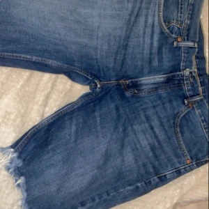 Levi's jeansshorts med rå kant - Säljer ett par klassiska blå jeansshorts från Levi's med rå, fransig kant nertill. Shortsen har fem fickor, gylf med dragkedja och knapp samt den ikoniska röda Levi's-lappen på bakfickan. Perfekta för en avslappnad stil.