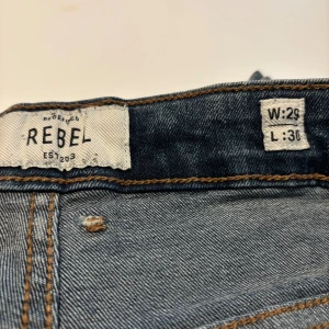 Blå distressed jeans från Rebel W29 L30 - Snygga blå jeans från Redefined Rebel med slitna detaljer och distressed look. Klassisk femficksmodell med ljus tvätt och coola slitningar på benen. Perfekt för dig som gillar en urban och avslappnad stil.