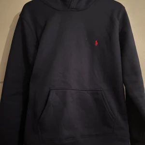 Mörkblå hoodie från Polo Ralph Lauren - Mörkblå hoodie från Polo Ralph Lauren med klassisk röd logga broderad på bröstet. Tröjan har huva, magficka och är långärmad. Perfekt för dig som gillar stilren och enkel design. Skriv för fler frågor 