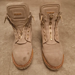 Beige mockaboots från Balmain - Snygga beige boots från Balmain i mjuk mocka med grov sula och detaljer i guld. Skorna har snörning och dragkedja framtill, samt Balmain-logga på plös och sula. Perfekt chunky look med rund tå och robust yttersula. Nypris:15000-25000 