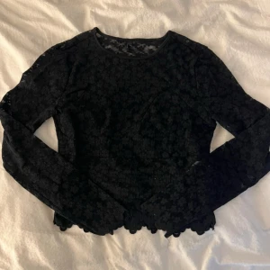 Svart blommig spetsblus med lång ärm - Säljer en svart blus i spets med blommigt mönster och lång ärm. Toppen har en rund halsringning och är lätt transparent med fina detaljer längs nederkanten. Perfekt för dig som gillar en feminin och edgy stil. Använd fåtal gånger 