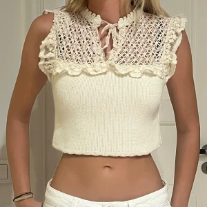 Cremefärgad stickad topp med spets från Zara - Supersöt cremefärgad stickad topp från Zara med croppad passform. Toppen har ett ribbat liv och detaljerad spets med volang upptill samt knytning vid halsen. Ärmlös design med romantiska virkade partier som ger en unik look. Perfekt för dig som gillar feminina detaljer.