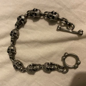 Silverfärgat armband med dödskallar - Coolt armband i silverfärg med detaljerade dödskallar som länkar och ett unikt benformat spänne. Perfekt för dig som gillar edgy och alternativ stil. Armbandet har en robust känsla och sticker verkligen ut.