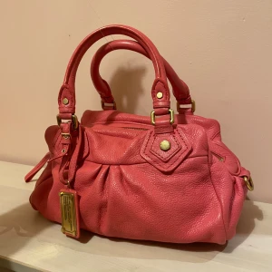 Rosa handväska från Marc by Marc Jacobs - Säljer en snygg rosa handväska från Marc by Marc Jacobs i mjukt skinn med guldfärgade metalldetaljer. Väskan har dubbla handtag, avtagbar axelrem och dekorativa nitar. Perfekt storlek för det viktigaste och har coola detaljer som ger extra stilpoäng.