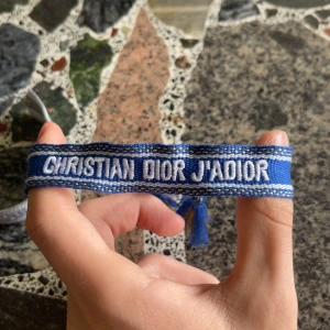 Blå Christian Dior J'Adior armband - Snyggt blått armband från Christian Dior med texten 'J'Adior' broderad i vitt. Armbandet har dekorativa tofsar i blått och guld och är gjort i ett mjukt, vävt material. Perfekt för dig som vill ha en cool designeraccessoar med statement.