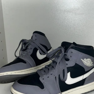 Nike Air Jordan 1 Mid Grå - Säljer ett par Nike Air Jordan 1 Mid i grått och marinblått med vita detaljer och klassisk swoosh. Skorna har snörning, rund tå och platt sula. Tillverkade i skinn och syntetmaterial, perfekta för dig som vill ha en ikonisk sneaker med streetkänsla.