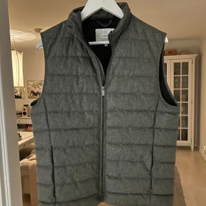 Grå quiltad väst - Snygg grå dunväst från Bläck med quiltad design och rak passform. Västen har ståkrage och dragkedja framtill, perfekt att slänga över en hoodie eller tröja. Materialet är mjukt och lätt, vilket gör den enkel att bära under kyliga dagar.