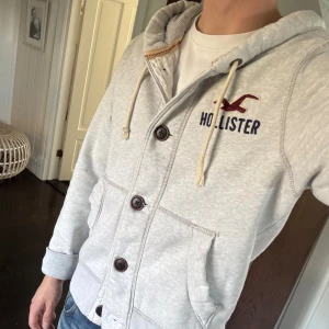Hollister Hoodie  - Snygg grå Hollister Hoodie med knappar på framsidan och loggan på bröstet. Riktigt bra skick. Storlek: Small. Passar perfekt nu till hösten!