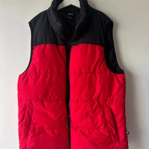 Röd och svart dunväst från Cropp - Snygg dunväst från Cropp i rött och svart med hög krage och dragkedja framtill. Västen har en quiltad look och två fickor på sidorna. 