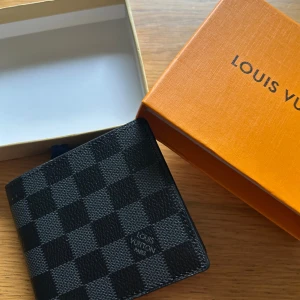 Louis Vuitton svart rutig plånbok - Snygg svart plånbok från Louis Vuitton med klassiskt rutigt Damier-mönster. Plånboken är tillverkad i exklusivt skinn och har flera fack för kort och sedlar. Perfekt storlek för att ha med sig överallt och levereras med original ask.