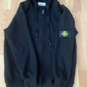 Svart hoodie från Stone Island - Svart hoodie från Stone Island med dragkedja framtill och klassisk logga på vänster ärm. Tröjan har huva med snörning och ribbade muddar. Perfekt för dig som gillar streetwear och vill ha en stilren look.