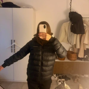 Svart pufferjacka med huva - Säljer en svart pufferjacka från Peak Performance med huva och dragkedja framtill. Jackan har en quiltad design och långa ärmar med muddar. Perfekt för dig som vill ha en varm och stilren jacka med clean look.💗