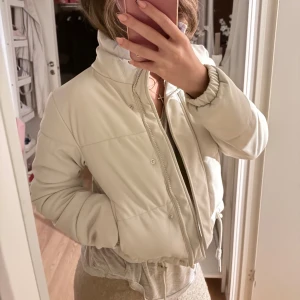 Créme färg croppad pufferjacka - Säljer en snygg croppad pufferjacka i creme färg med hög krage och dragkedja framtill. Jackan har långa ärmar, elastiska muddar och snörning i nederkant för en cool passform. Storlek XS använt 2 gånger 