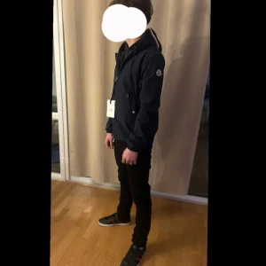 Mörkblå vindjacka från Moncler - Snygg mörkblå vindjacka från Moncler  i storlek 1=S med huva och snörning. Jackan har dragkedja, Moncler-logga på ärmen och en cool rödvit detalj på insidan av huvan. Perfekt för dig som gillar stilrena och sportiga plagg.