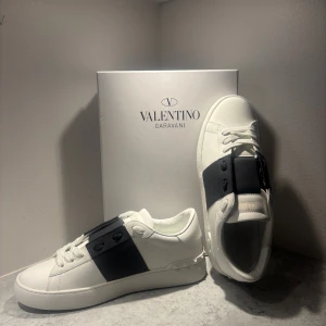 Valentino Garavani Open sneakers vit/svart/grå - Säljer ett par snygga Valentino Garavani Open sneakers i vitt skinn med bred svart läderdetalj över snörningen. Klassisk rund tå, platt sula och diskreta nitar baktill. Lyxig känsla och stilren design, perfekt för dig som gillar exklusiva sneakers. Hör av vid fundering pris kan sänkas vid snabb affär.