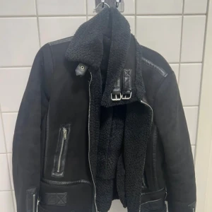 Svart teddyjacka från Hailys - Svart teddyjacka från Hailys med feta detaljer i skinnimitation, bred krage och dragkedjor på fickorna. Jackan har en schysst oversized look, mjukt teddyfoder och coola spännen vid halsen. Perfekt för dig som vill ha en varm och stilren mocka jacka.