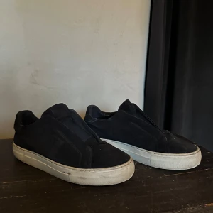 Arigato skor - Blåa sneakers från Axel Arigato med stilren design och platt vit sula. Skorna har små slitningar inne. Annars är dom bara smutsiga. Skorna är tillverkade i mocka och har en låg profil utan synlig snörning för en clean look. Perfekta för dig som gillar minimalistisk stil och vill ha något som funkar till jeans eller dressade byxor. Pris kan diskuteras men inte för lågt