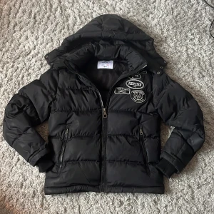 Svart pufferjacka från Mercier - Svart pufferjacka från Mercier med huva och coola patches på bröstet. Jackan har dragkedja framtill, två fickor med dragkedja och ribbade muddar vid ärmsluten. Perfekt för kalla dagar och riktigt snygg streetstil.