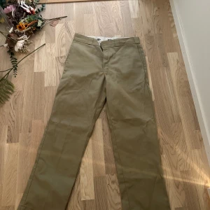 Dickies 874 khakifärgade chinos - Klassiska Dickies 874 Original Fit chinos i khakigrönt. Byxorna har rak passform, bälteshällor och två bakfickor, varav en med knapp. Tillverkade i slitstark bomullsblandning med dragkedja och knapp framtill. Perfekta för en clean och tidlös stil.
