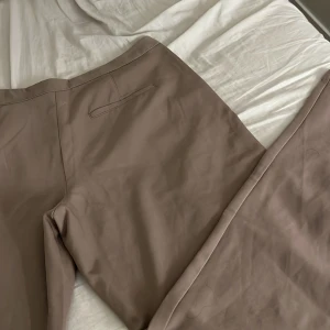 Beige wide kostymbyxor - Snygga beige kostymbyxor med breda ben och pressveck framtill. Byxorna har diskreta fickor och en stilren look som passar perfekt till en trendig outfit. Materialet känns mjukt och har en lätt glans, vilket ger en exklusiv känsla. 