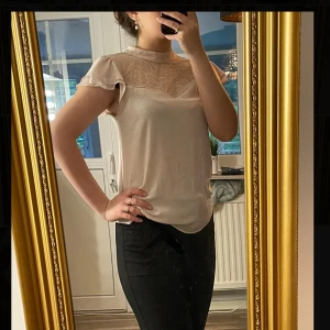 Beige blus med spetsdetalj - Säljer en beige blus med korta ärmar och hög hals. Överdelen har en snygg spetsdetalj. Blusen är lätt och har en mjuk, flowy passform. 