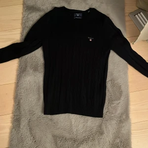 Mörkblå  kabelstickad tröja från Gant - Mörkblå kabelstickad tröja från Gant i premium bomull. Klassisk rund halsringning, logga på bröstet och ribbade muddar. Perfekt för dig som gillar stilrena och tidlösa plagg med snygg passform, ny pris 1600