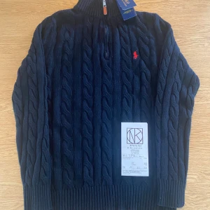 Mörkblå kabelstickad tröja Polo Ralph Lauren - Kabelstickad mörkblå tröja från Polo Ralph Lauren med halv dragkedja och hög krage. Klassisk röd broderad logga på bröstet. Tröjan är i ull och har ribbade muddar och nederkant. Perfekt för dig som gillar preppy och stilren look.