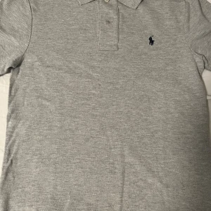 Polo Ralph Lauren piké - Snygg grå pikétröja från Polo Ralph Lauren i slim fit-modell. Tröjan har klassisk krage, knappar framtill och den ikoniska broderade loggan på bröstet. Tillverkad i mjuk bomull som känns skön mot huden. Perfekt för en clean och stilren look.