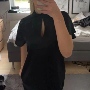 Svart blus - Svart blus från b.young med hög hals och snygg öppning framtill. blusen har korta ärmar och en rak, enkel passform som känns både stilren och modern. Perfekt för dig som gillar minimalistisk stil och vill ha något som sticker ut med en cool detalj vid halsen.