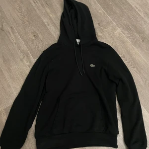 Svart hoodie från Lacoste med huva - Svart hoodie från Lacoste i klassisk passform med broderad krokodil på bröstet. Tröjan har huva med snörning , använd några gånger är som ny nästan jätte bra  skick.