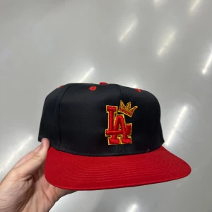 Svart och röd LA keps med krona - Snygg snapback keps i svart med röd skärm och broderad LA-logga i rött och gult, toppad med en gul krona. Kepsen har röda detaljer och justerbar baksida. Perfekt för dig som vill sticka ut med streetstyle.