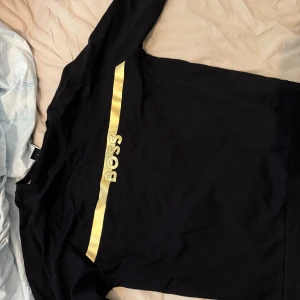 Svart sweatshirt BOSS - Svart långärmad tröja från BOSS med en bred, glansig guldfärgad rand och BOSS-logga över bröstet 