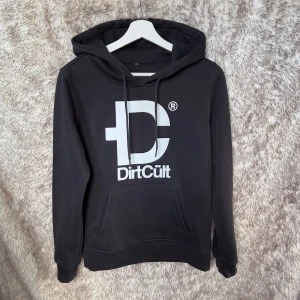 Svart hoodie från DirtCült i M - Svart hoodie från DirtCült med stor vit logga framtill. Klassisk modell med huva, snörning och känguruficka. Mjuk insida och ribbade muddar vid ärmslut och nederkant. 