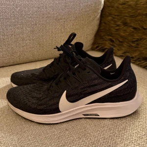 Svarta Nike Zoom sneakers - Säljer ett par svarta Nike Zoom sneakers med vit sula och stor vit swoosh på sidan. Skorna har snörning och en stickad ovandel i syntetmaterial med subtilt mönster. Perfekta för dig som gillar sportig stil och vill ha bekväma skor till träning eller häng.