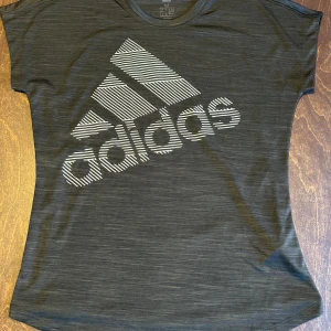 Adidas t-shirt - Svart t-shirt från Adidas med stor vit logga framtill i ett randigt mönster. T-shirten har korta ärmar och är tillverkad i ett mjukt, lätt material som passar perfekt för träning. Knappt använd 