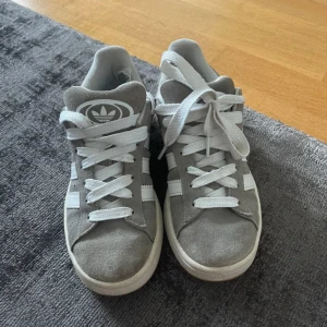 Grå Adidas Campus sneakers i mocka - Snygga Adidas Campus sneakers i grå mocka med vita detaljer och klassiska tre ränder på sidorna. Skorna har vit sula, platt gummisula och snörning framtill. Perfekta för en clean och stilren look. Adidas-logga på plösen och Campus-tryck på sidan. Storlek 37 1/3.