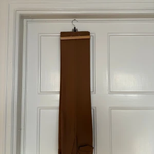 Bruna kostymbyxor med raka ben - Snygga bruna kostymbyxor med raka ben och klassiska pressveck. Byxorna har hällor för bälte och en bakficka med lock. Perfekta för en stilren look och passar till skjorta eller t-shirt. Mäter 34 cm rakt över midjan och 77 cm från gren till benslut. Färgen på första bilden stämmer bäst överens med verkligheten.
