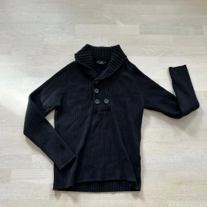 Svart ribbad kofta med knappar - Säljer en svart ribbad kofta med bred krage och  stora knappar framtill. Tröjan har långärmade och är tillverkad i ett stickat material som känns mjukt och skönt. Perfekt att slänga på sig när det är lite kyligt ute. 