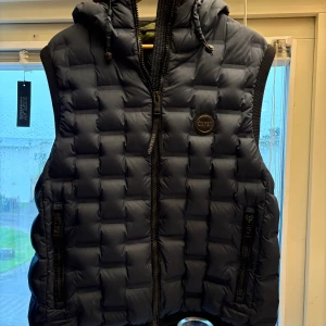 Mörkblå dunväst Colmar Research edition - Snygg mörkblå dunväst från Colmar med quiltad design och huva. Västen har dragkedja framtill, två fickor med dragkedja och coolt grönt foder på insidan. Köpt från NK gbg 2022 och kommer från Research släppet, storleken är M