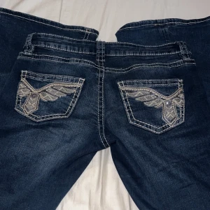 Mörkblå bootcut jeans med broderi - Snygga mörkblå jeans med bootcut passform och coola broderade detaljer på bakfickorna. Jeansen har kontrastsömmar och en klassisk femficksdesign. Perfekta för dig som vill sticka ut med detaljer och gillar en lite vidare benslut.