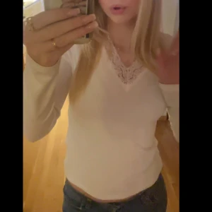 Vit långärmad topp med spets - Säljer en vit långärmad topp med v-ringning och snygg spetsdetalj vid halsen. Toppen har en figurnära passform och är perfekt att styla med jeans eller kjol. Superfin och enkel att matcha till olika looks!