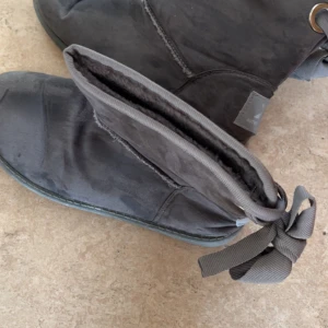 Grå boots från Xti med rosett - Säljer ett par grå boots från Xti med snygg rosettdetalj baktill. Skorna är i mockaimitation och har en platt sula med grovt mönster undertill. Perfekta för höst och vinter när du vill ha både stil och komfort.