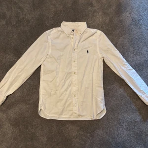 Vit slim fit skjorta från Polo Ralph Lauren - Vit skjorta från Polo Ralph Lauren i slim fit-modell. Klassisk design med knappar hela vägen, button-down krage och ikonisk broderad logga på bröstet. Långärmad och tillverkad i mjuk bomull som känns skön mot huden. Perfekt för dig som gillar stilrena och tidlösa plagg.