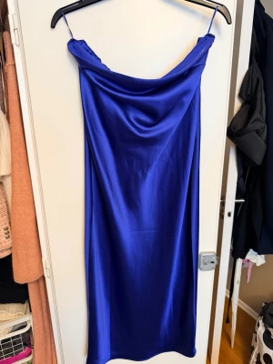 Blå satin klänning från GinaTricot  - Säljer en snygg blå midiklänning från GinaTricot i satin med axelbandslös design och draperad halsringning. Klänningen har ett band i ryggen som kan knytas och en mjuk, glansig finish som ger en lyxig känsla. Perfekt för dig som vill sticka ut med färg.