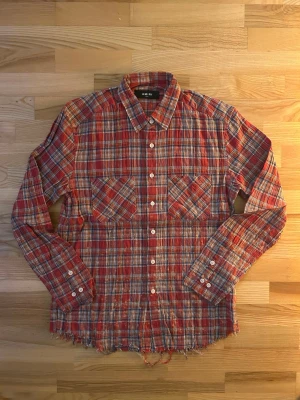 Rutig röd skjorta från Amiri - Säljer en rutig skjorta från Amiri i rött, blått och vitt med stora broderade bokstäver på ryggen. Skjortan har långa ärmar, två bröstfickor och knappar framtill. Materialet är bomull med lite elastan och nederkanten är rå och fransig för en edgy vibe.