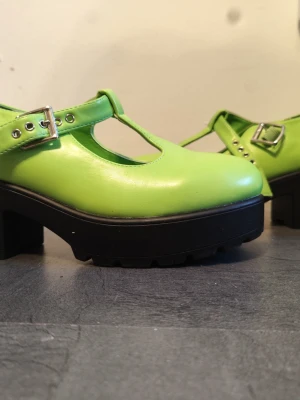 Limegröna Mary Jane-plattformsskor från KOI x tellietubbies  - Säljer ett par limegröna Mary Jane-plattformsskor från KOI x tellietubbies med chunky svart sula och bred klack. Skorna har rund tå, T-rem med silvriga spännen och är tillverkade i syntetmaterial. Perfekt statement-piece för dig som gillar färg och coola detaljer. Använda ca två gånger 