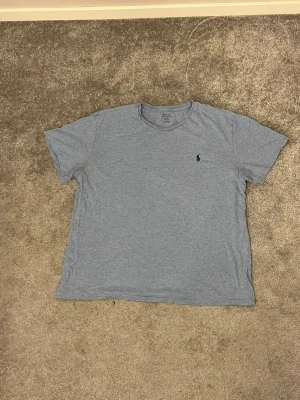 Ljusblå t-shirt från Polo Ralph Lauren - Säljer en ljusblå t-shirt från Polo Ralph Lauren med klassisk rund hals och korta ärmar. T-shirten har en liten mörkblå Polo-logga broderad på bröstet. Materialet är mjuk bomull som känns skönt mot huden. Perfekt för dig som gillar stilrena och enkla plagg. Tröjan är använd så slitage finns 