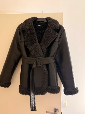 Svart teddyjacka med bälte Gina Tricot - Svart teddyjacka från Gina Tricot i storlek S. Jackan har fluffig teddykrage och muddar, samt ett snyggt bälte i midjan. Materialet är skinnimitation med mjuk teddy på insidan och detaljerna ger en cool vibe. Perfekt för kalla dagar när du vill vara både varm och stilren.